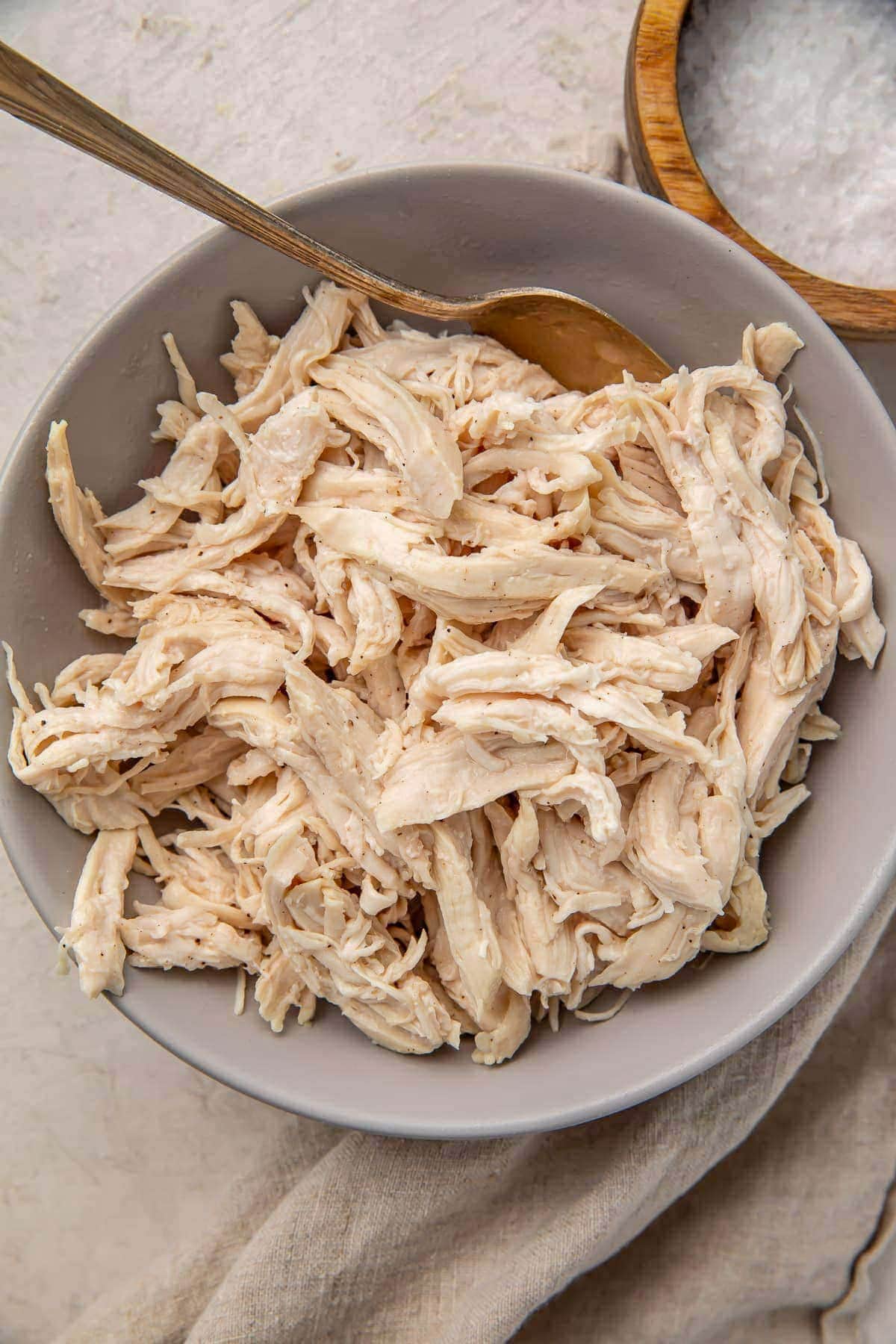 A light grey bowl holding large pieces of juicy shredded chicken with a utensil.