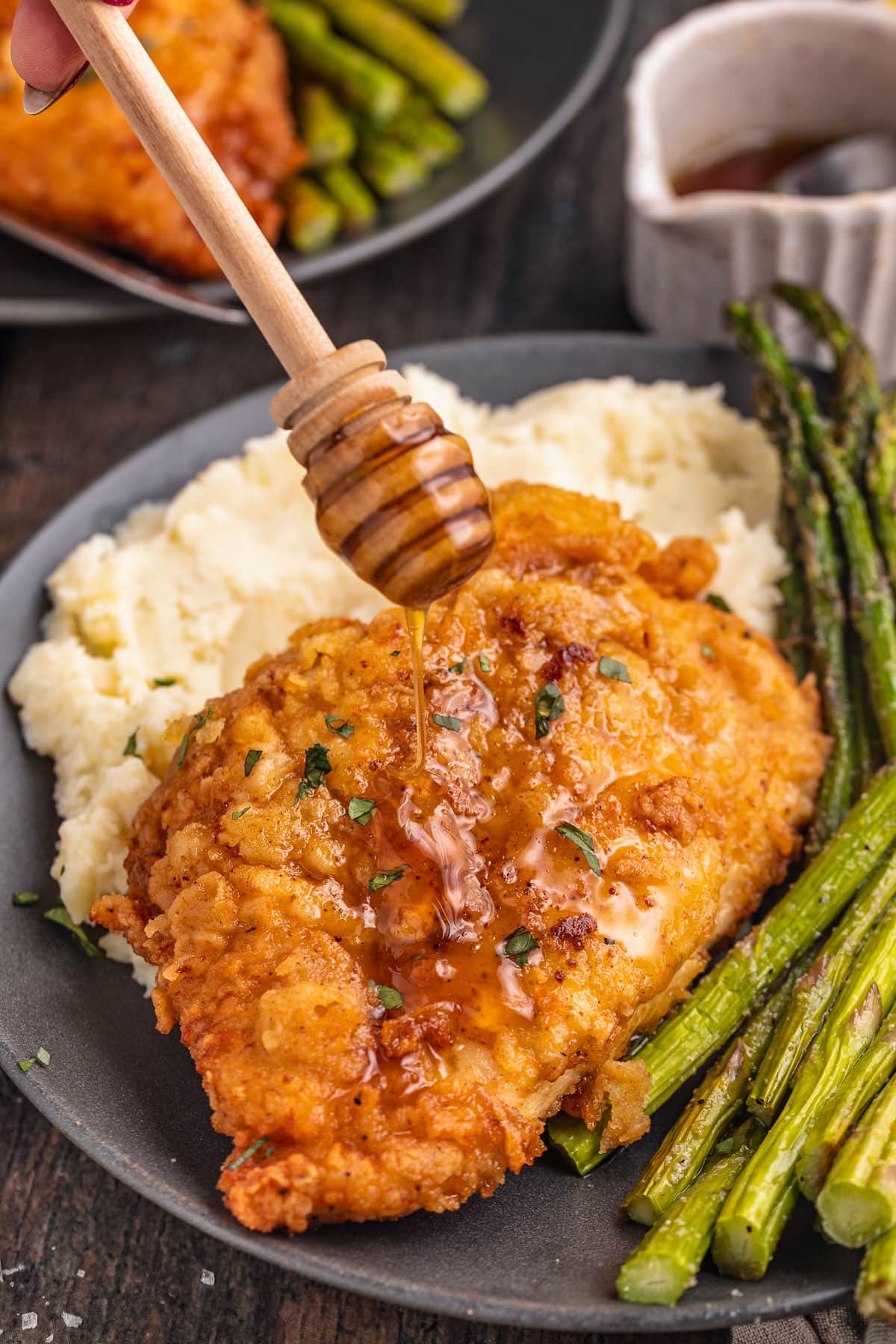 Crispy breaded chicken breast being drizzled with truffle honey on a plate next to asparagus and mashed potatoes.