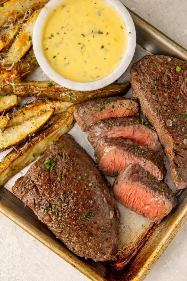 Medium rare steak and thick cut fries with a bowl of bearnaise sauce on a sheet pan