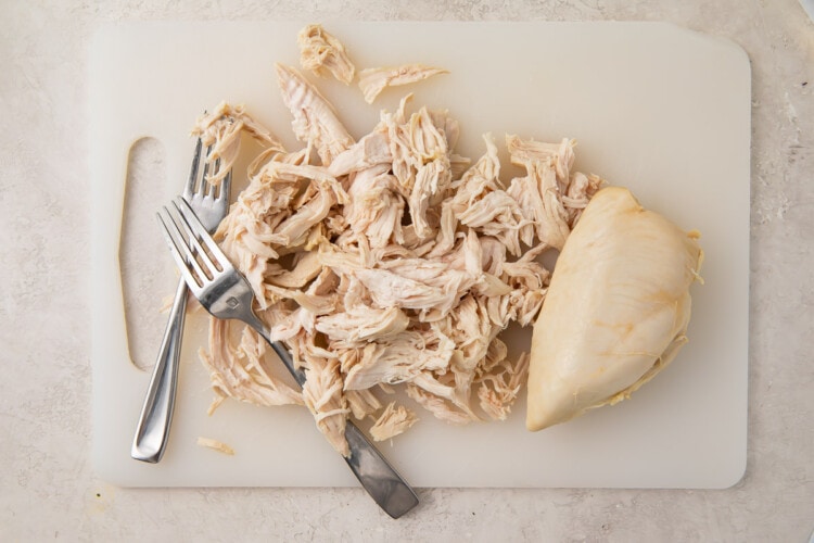 Shredded chicken breasts on a cutting board with 2 forks, next to whole cooked chicken breast
