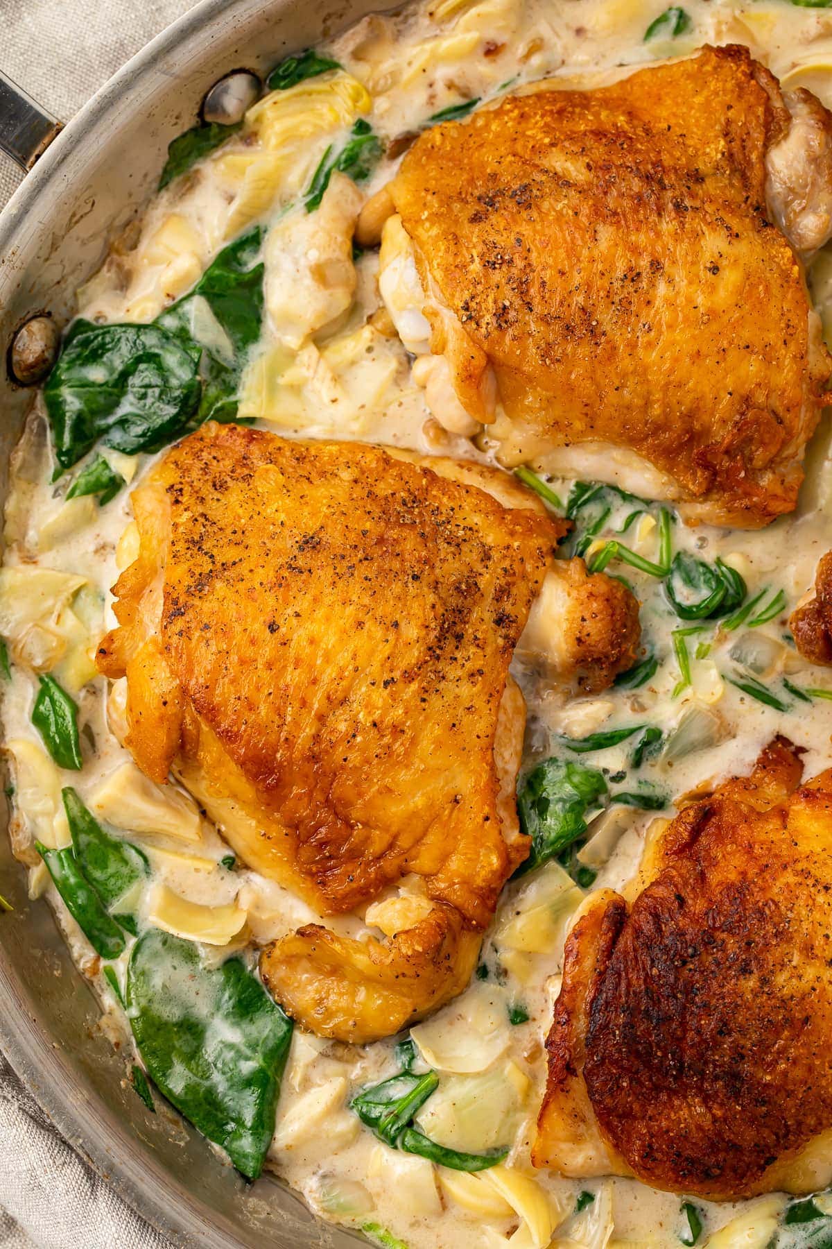 Close up photo of paleo spinach and artichoke chicken in a large silver pan. Chicken is uncovered by spinach artichoke sauce