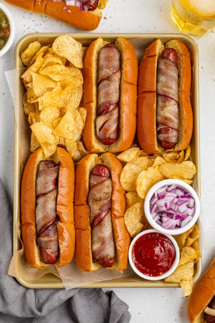 bacon wrapped hot dogs on a tray with chips, ketchup, and red onion
