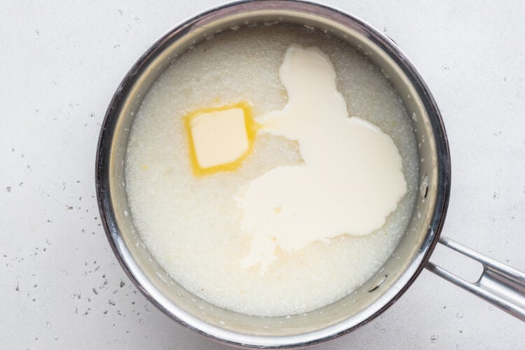 Overhead view of grits, water, butter, and heavy cream in a large silver saucepan.