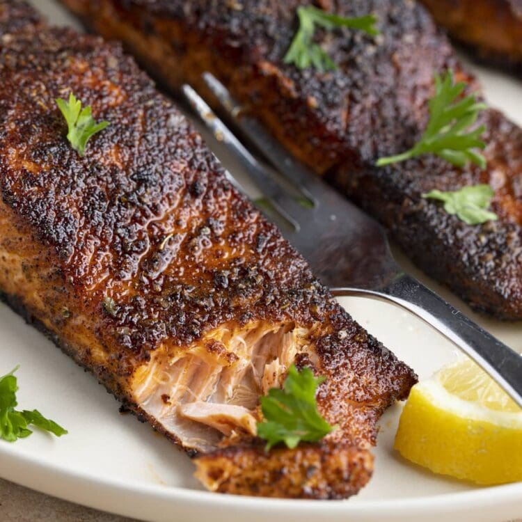 Angled view of blackened salmon on a white plate. The corner of the salmon has been cut into with a fork.