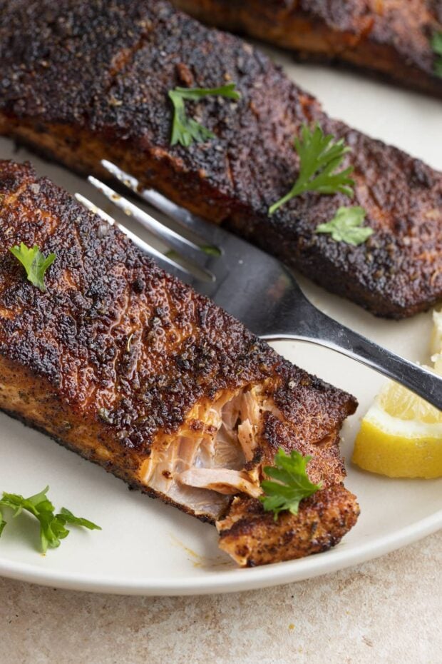 Angled view of blackened salmon on a white plate. The corner of the salmon has been cut into with a fork.