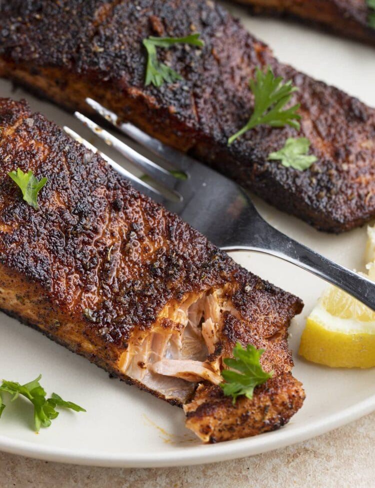 Angled view of blackened salmon on a white plate. The corner of the salmon has been cut into with a fork.