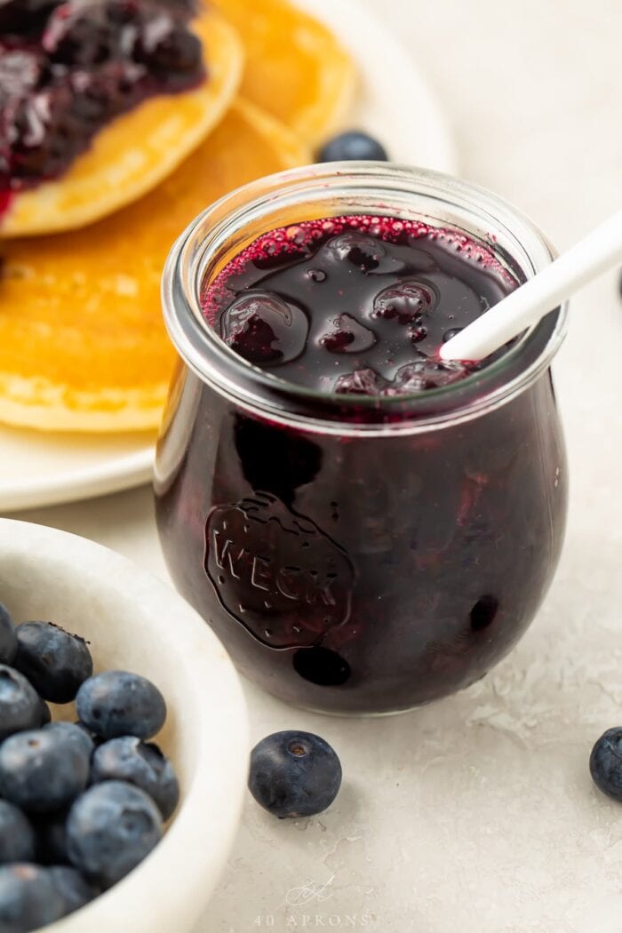 A clear glass jar of blueberry compote with a white spoon sticking out of the top. In the background is the edge of a plate of pancakes topped with blueberry compote. In the foreground is a small white bowl of blueberries