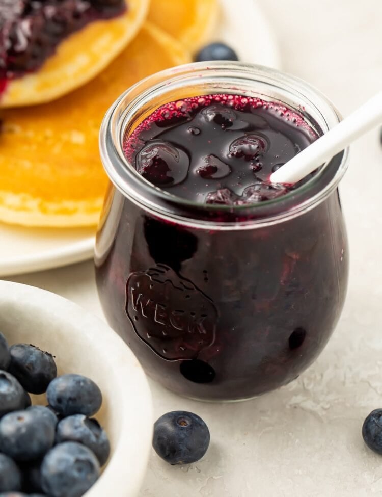A clear glass jar of blueberry compote with a white spoon sticking out of the top. In the background is the edge of a plate of pancakes topped with blueberry compote. In the foreground is a small white bowl of blueberries