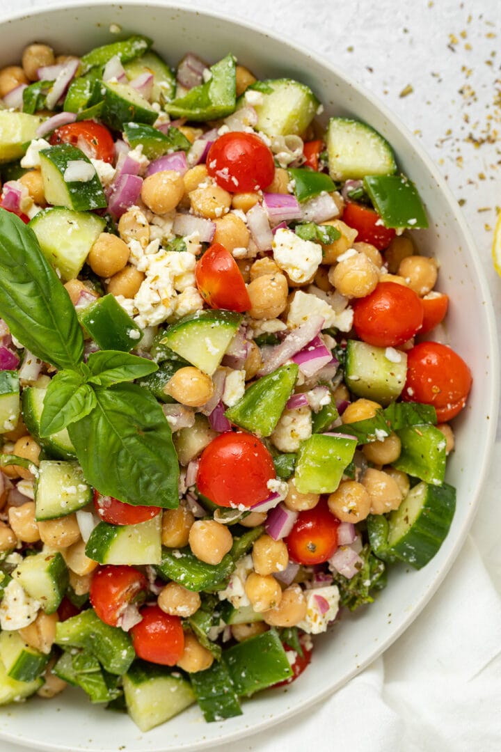 A large white bowl holding a Mediterranean-style chickpea salad with feta, bright red cherry tomatoes, chickpeas, cucumber, onion, and bell pepper.