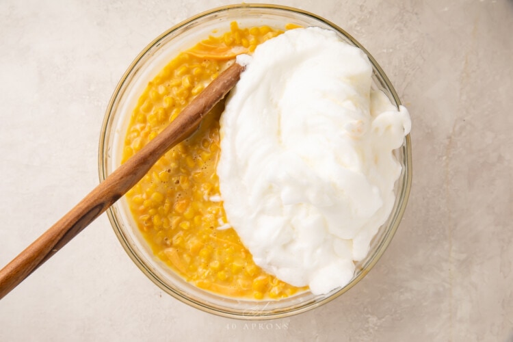 Mixture for corn souffle with whipped egg whites in a large glass bowl with a wooden spoon