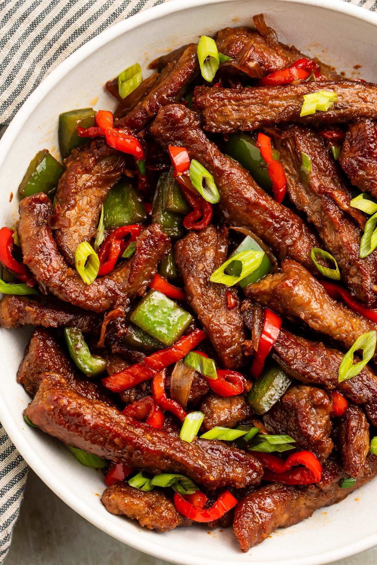 A large white bowl holding strips of hunan beef with vibrant red peppers and green onions.