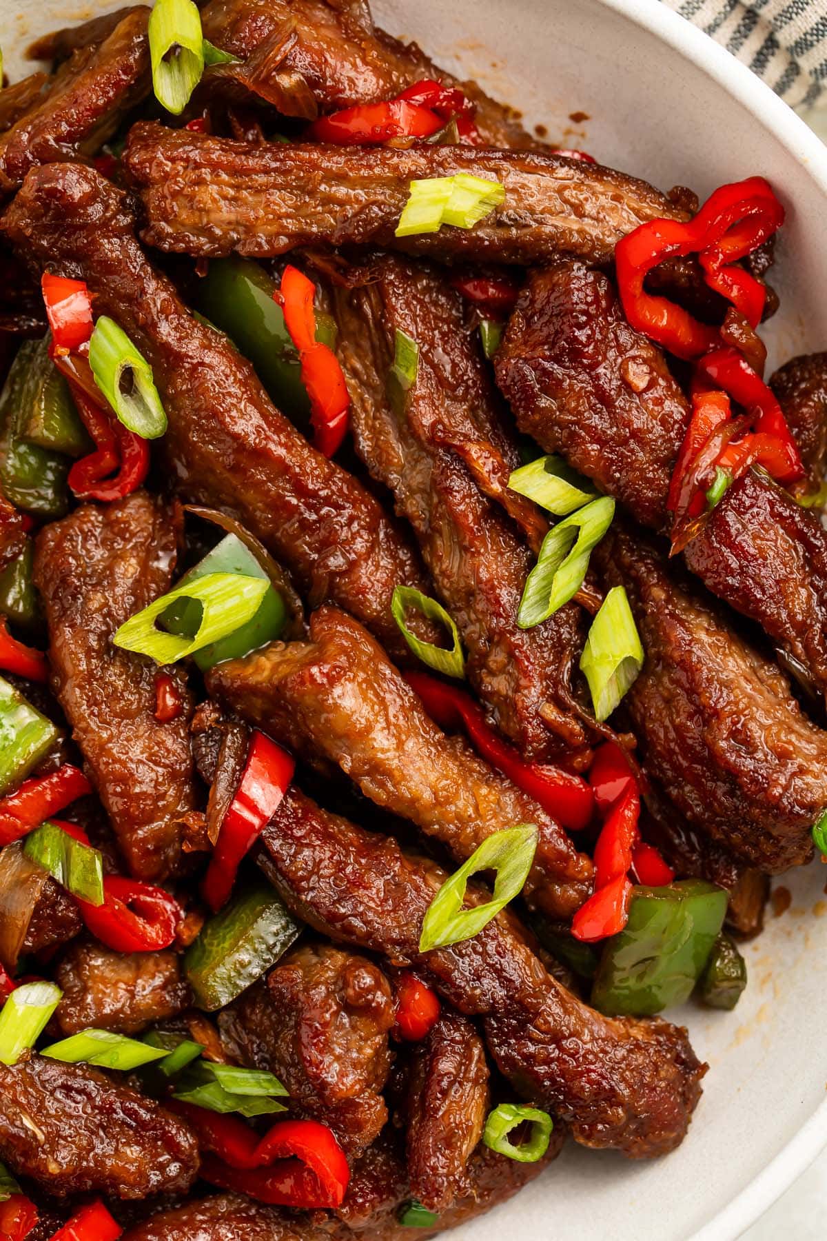 A large white bowl holding strips of hunan beef with vibrant red peppers and green onions.