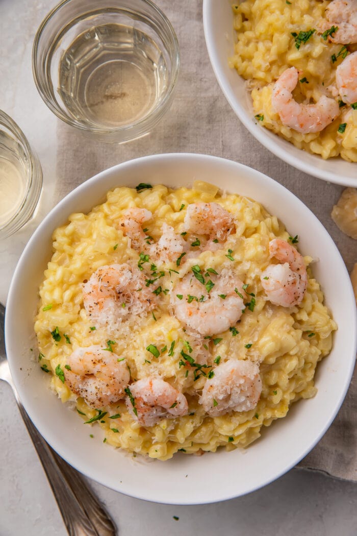 Zoomed out photo, taken from above, shows a bowl of shrimp risotto on a table next to a cloth napkin, with another bowl of shrimp risotto peeking in from the upper right corner