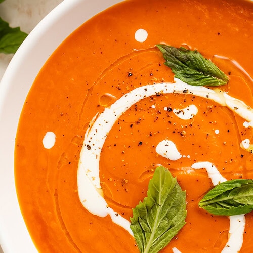Close-up overhead view of tomato bisque in a white bowl with a cream swirl and fresh basil leaves