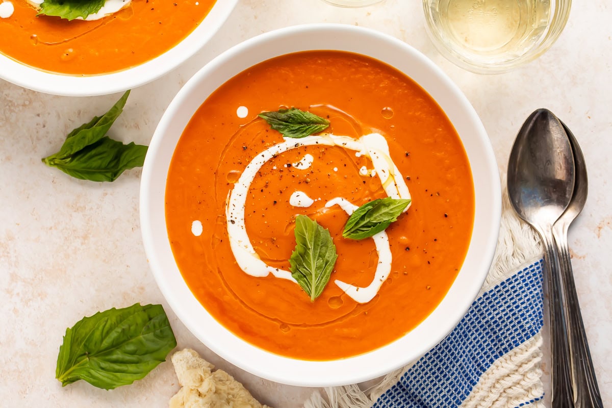 Overhead view of two white bowls of tomato bisque with cream swirls, basil, spoons, and crusty bread