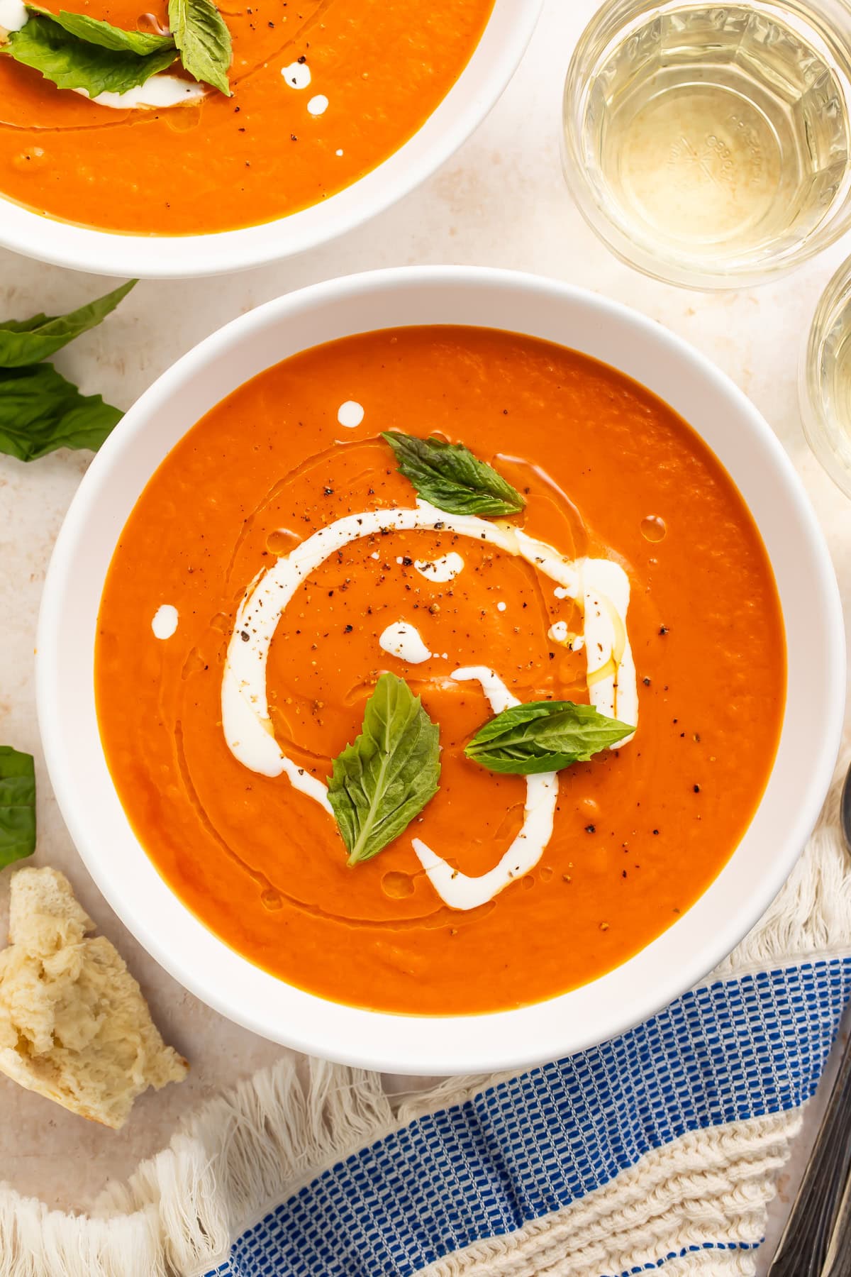 Overhead view of a white bowl of creamy tomato bisque with a cream swirl, fresh basil, and bread nearby