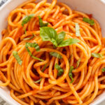 Close-up, overhead view of a white bowl of spaghetti swirled with red pomodoro sauce and topped with fresh basil leaves.