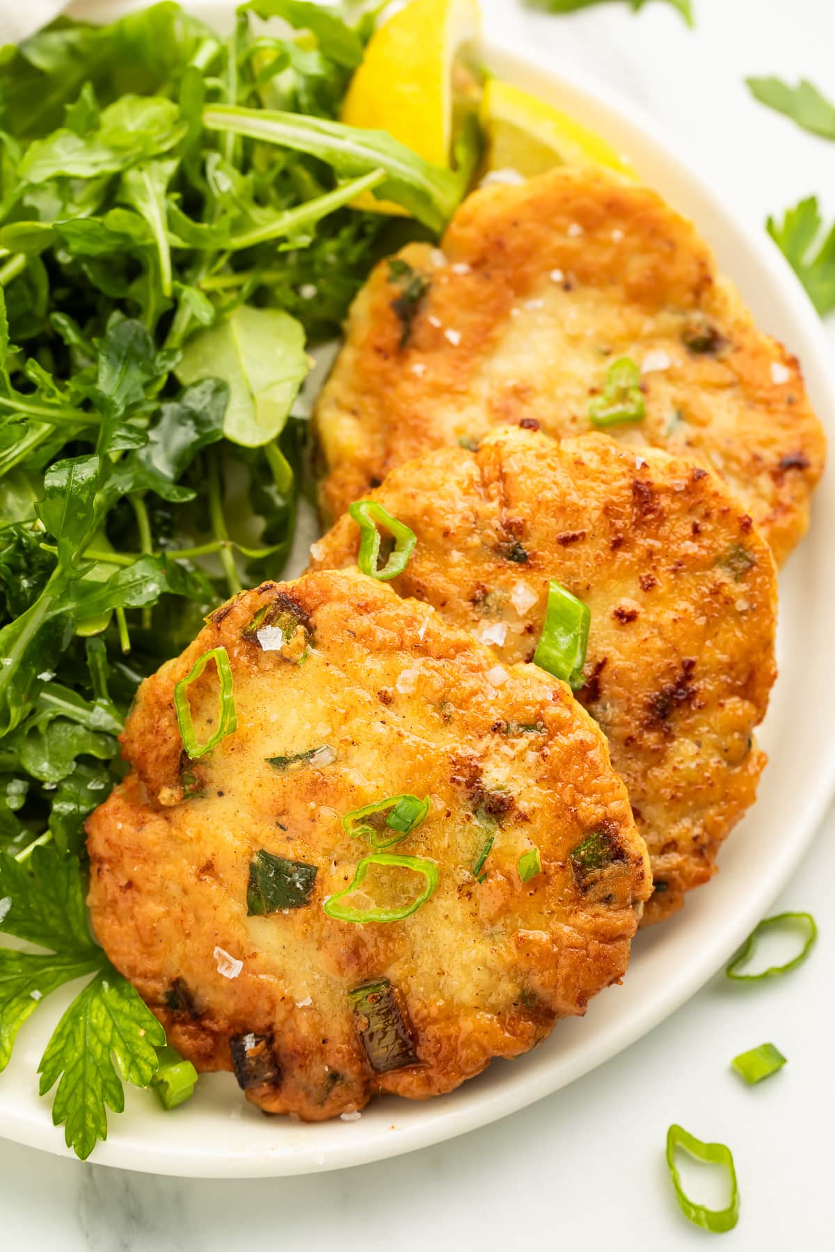 Three pan-fried, golden chicken patties arranged on a white plate with a leafy green side salad.
