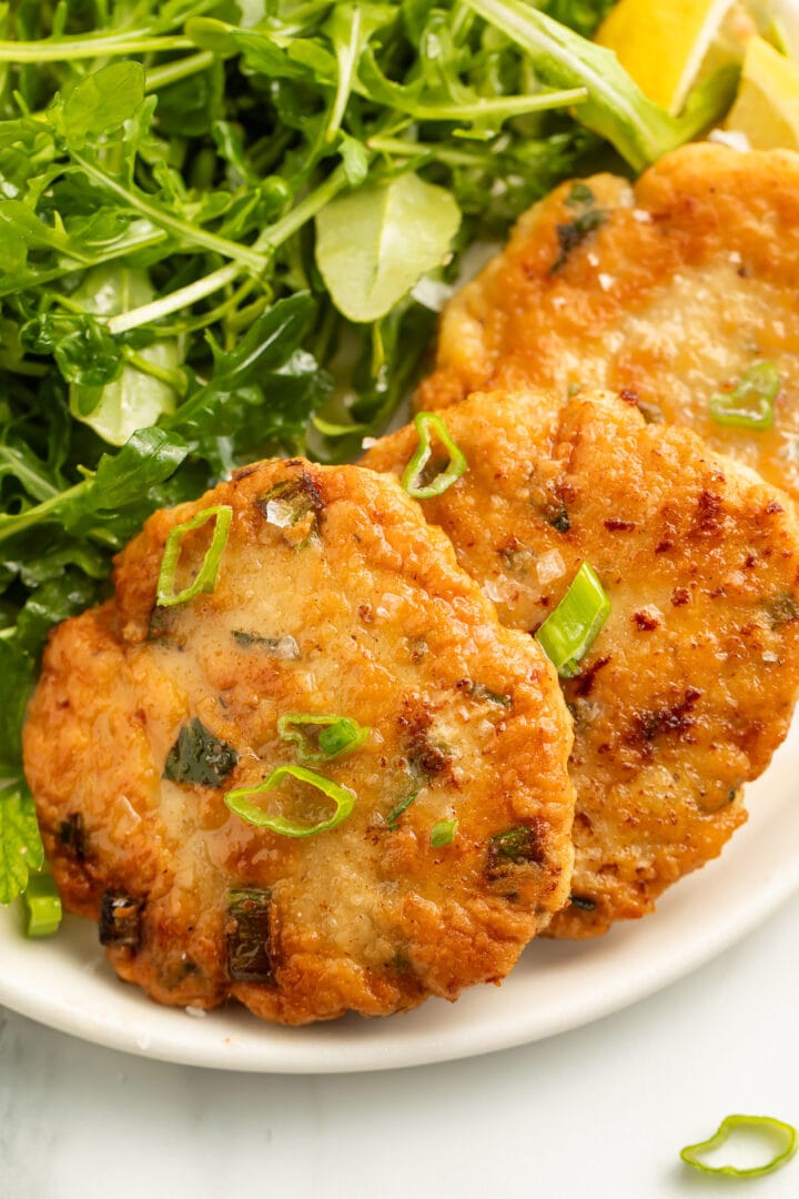 Three pan-fried, golden chicken patties arranged on a white plate with a leafy green side salad.
