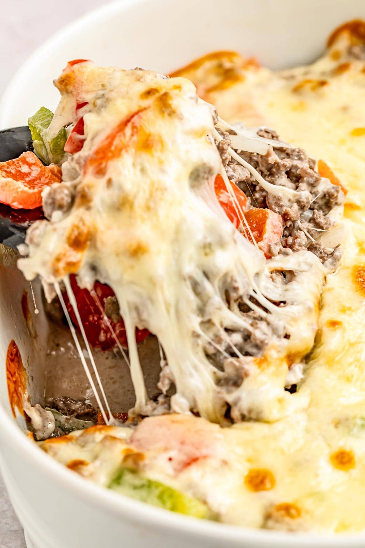 Extreme close-up of a portion of a Philly cheesesteak casserole with melty cheese, bell peppers, and ground meat being lifted out of a casserole dish with a spatula.