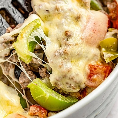 Extreme close-up of a spatula resting in a large casserole dish containing a Philly cheesesteak casseroel with melty cheese, bell peppers, and ground meat.