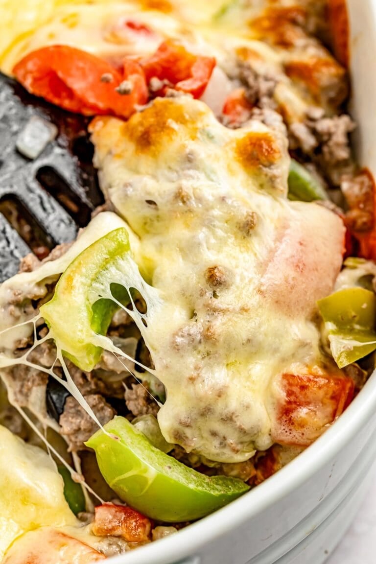 Extreme close-up of a spatula resting in a large casserole dish containing a Philly cheesesteak casseroel with melty cheese, bell peppers, and ground meat.