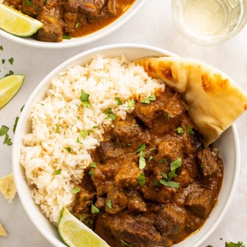 Two bowls of beef curry with white rice and naan on a white countertop.
