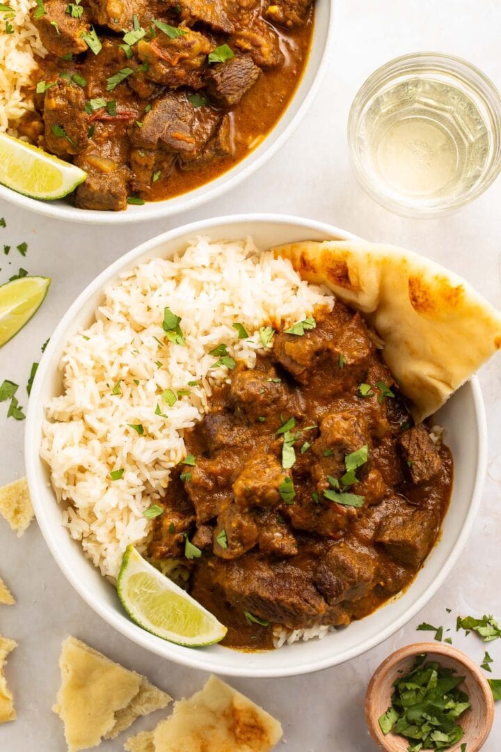 Two bowls of beef curry with white rice and naan on a white countertop.