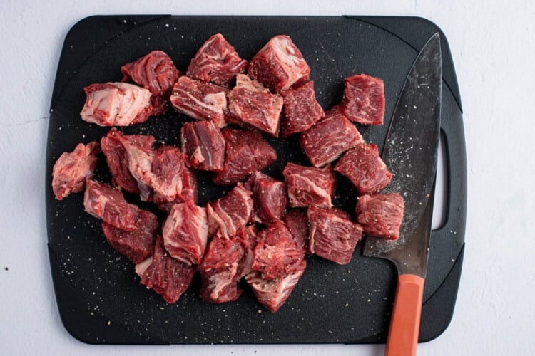 Chunks of beef seasoned with salt and pepper on a black cutting board with a large knife