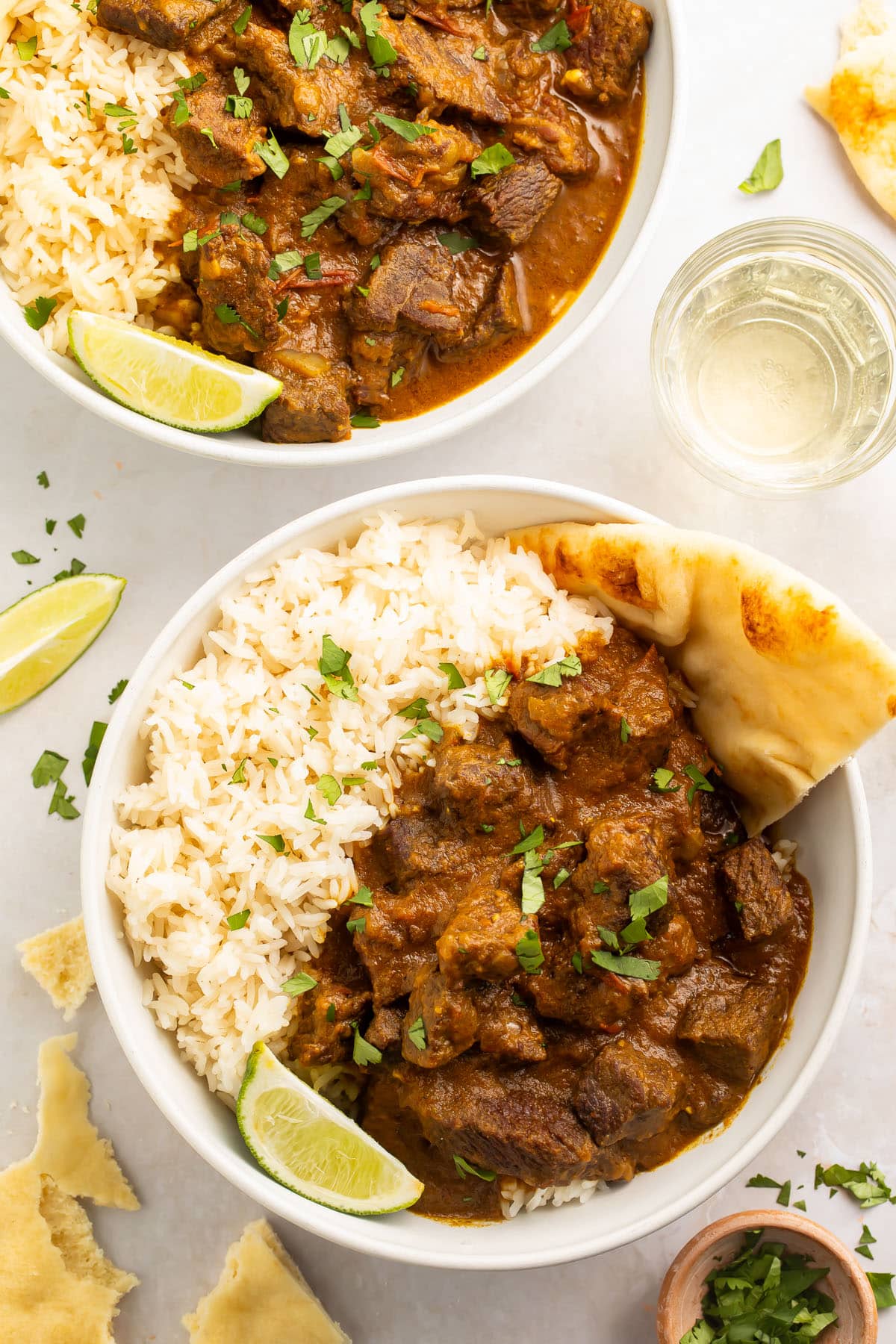 Two bowls of beef curry with white rice and naan on a white countertop.