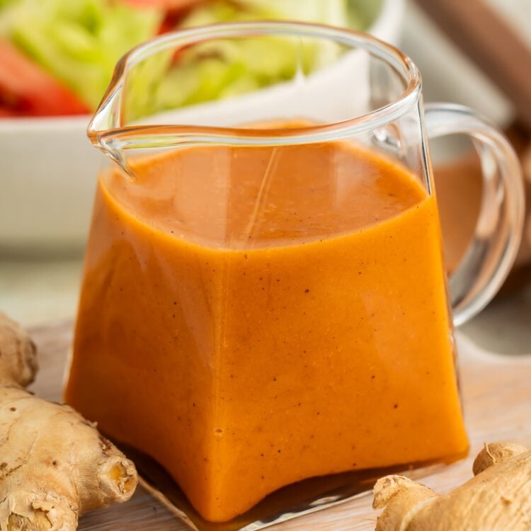 Orange-colored creamy ginger salad dressing in a small clear glass pitcher on a table in front of a bowl of salad.