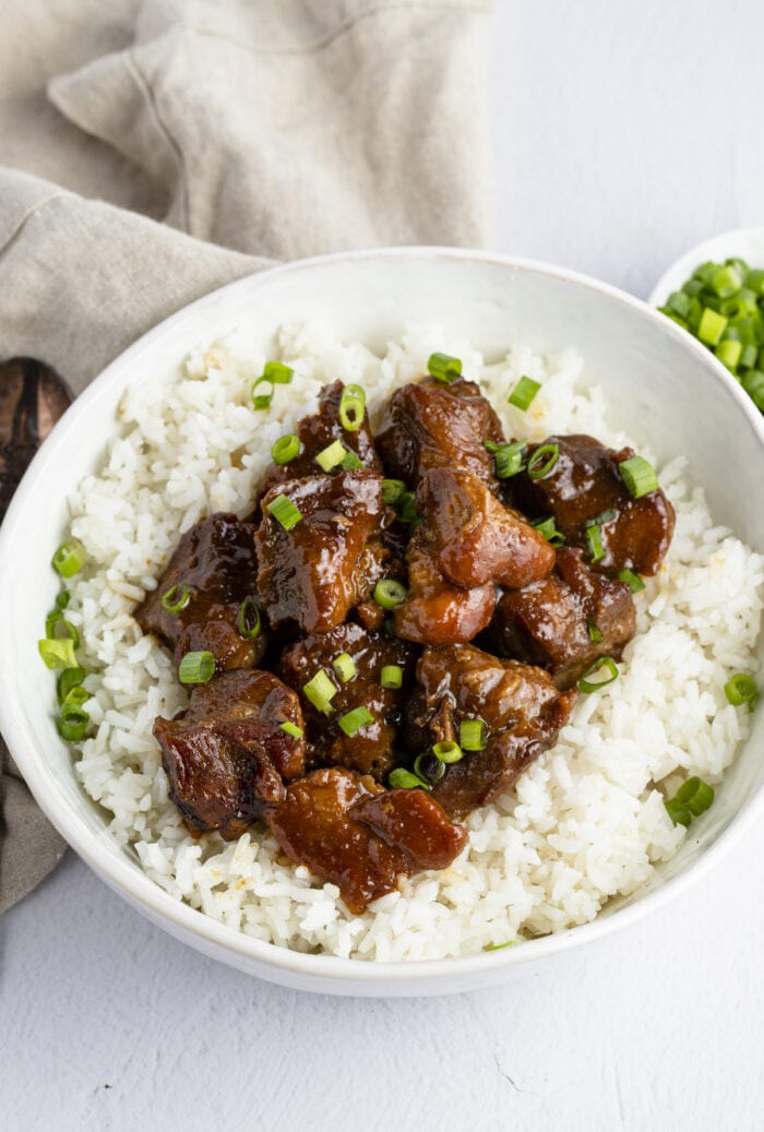 Overhead angled view of pork adobo on top of white rice in a bowl, with a napkin and a small bowl of chopped green onions in the background