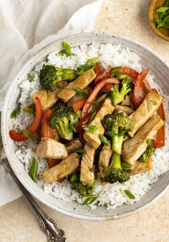 Pork stir fry on a bed of white rice in a white bowl with a napkin and spoon off to the side