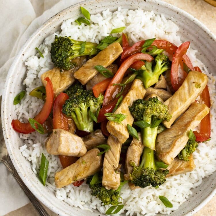 Pork stir fry on a bed of white rice in a white bowl with a napkin and spoon off to the side