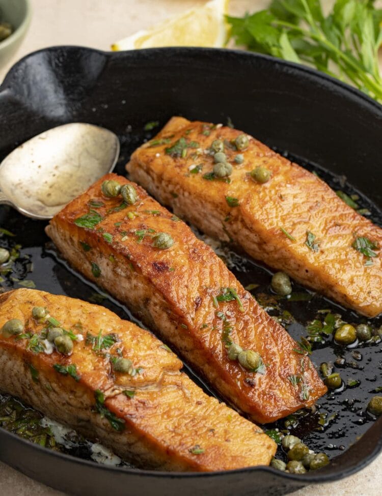 3 salmon meuniere fillets in a cast iron skillet with parsley and lemon wedges in the background