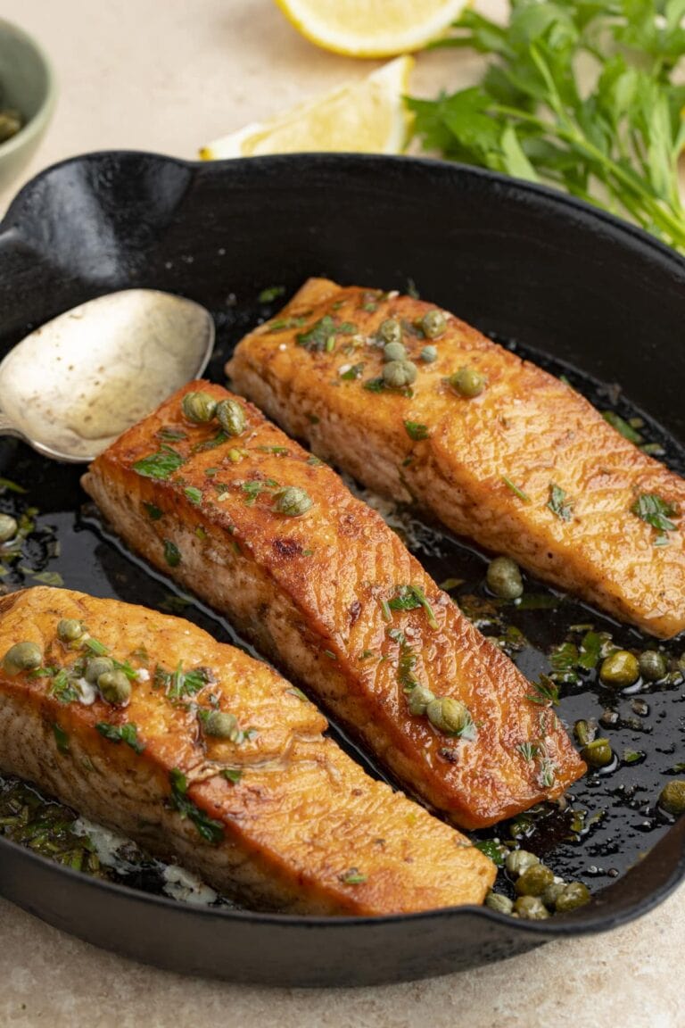 3 salmon meuniere fillets in a cast iron skillet with parsley and lemon wedges in the background