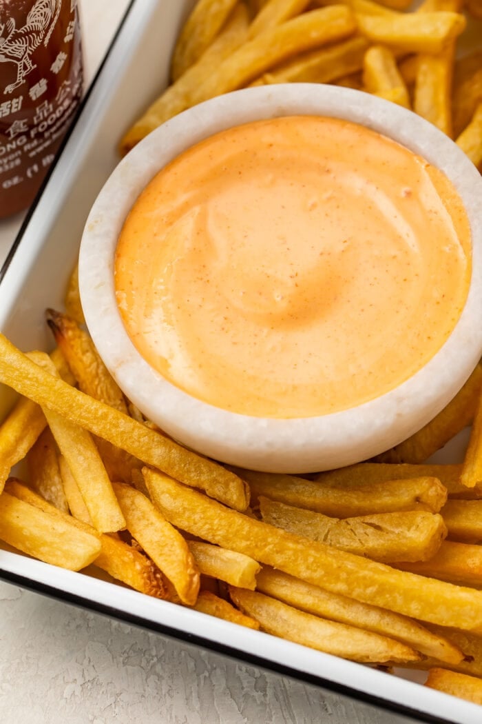 A small bowl of sriracha mayo on a baking sheet surrounded by french fries