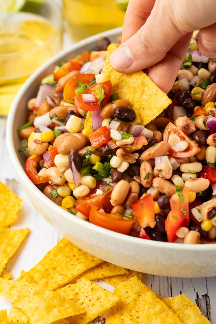 A white woman's hand holds a yellow corn tortilla chip as she uses it to scoop Texas caviar (cowboy caviar) out of a large white bowl.