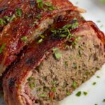 Bacon wrapped meatloaf slices on a white plate with ketchup and garnish