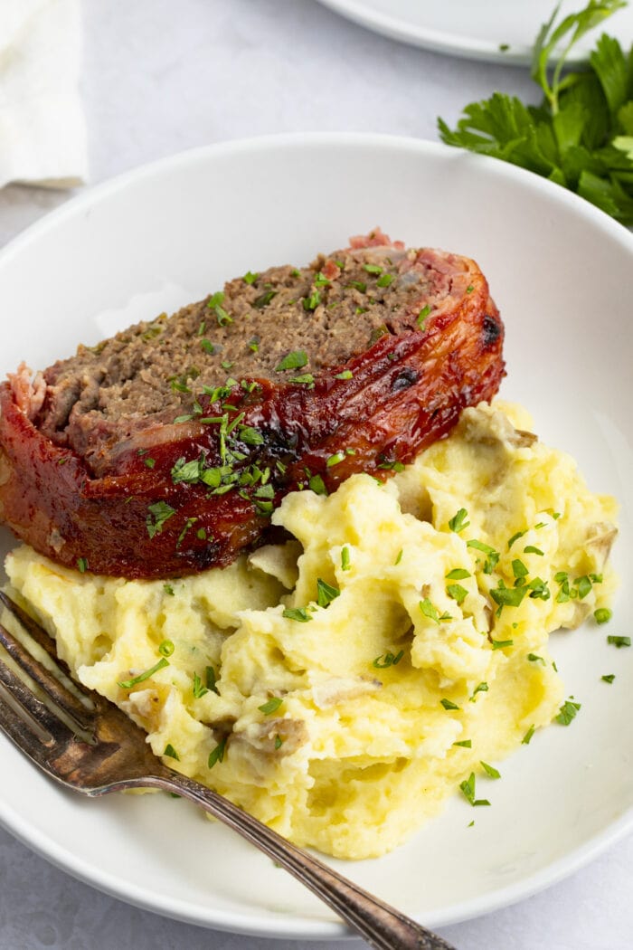 Bacon wrapped meatloaf on a bed of mashed potatoes on a white plate