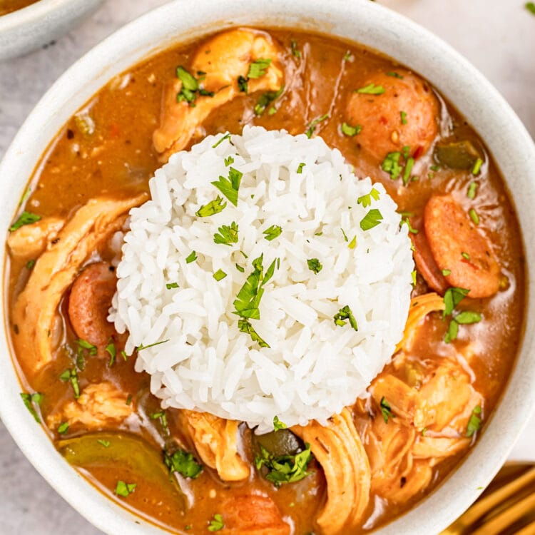 Overhead image of a white bowl holding deep reddish brown chicken an sausage gumbo with a mound of white rice in the center.