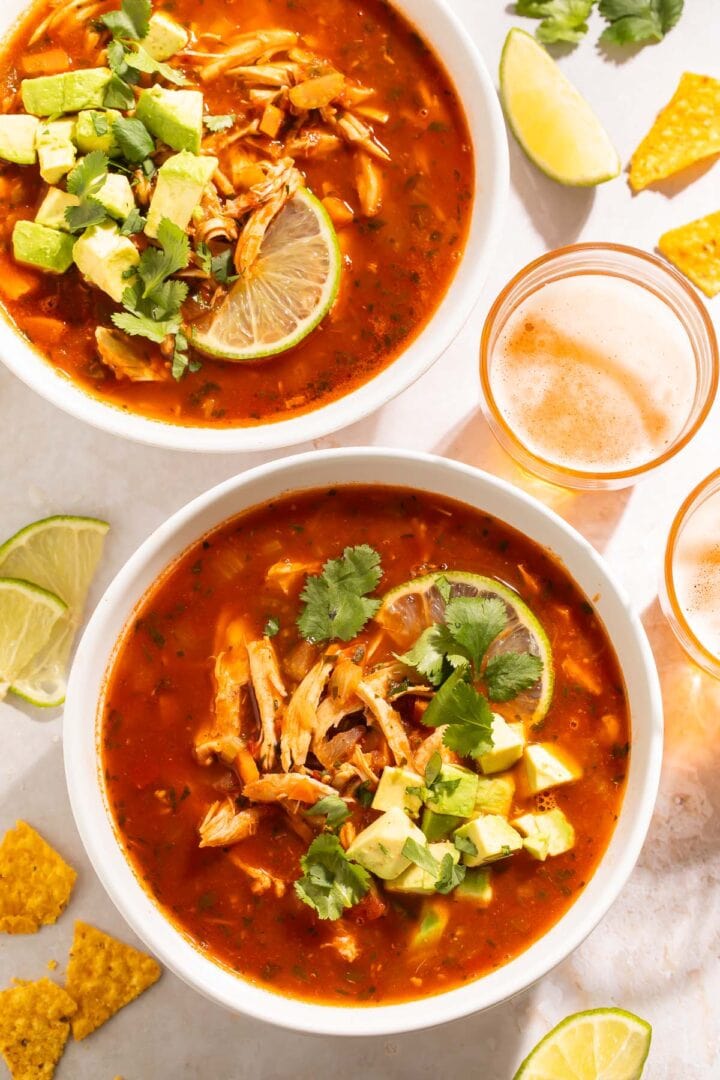 Two bowls of Mexican chicken soup on a table, surrounded by lime wedges, tortillas, and glasses of beer.