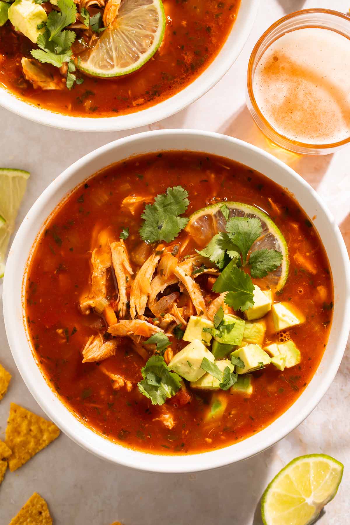 Close-up of a bowl of Mexican chicken soup on a table, surrounded by lime wedges, tortillas, and glasses of beer.