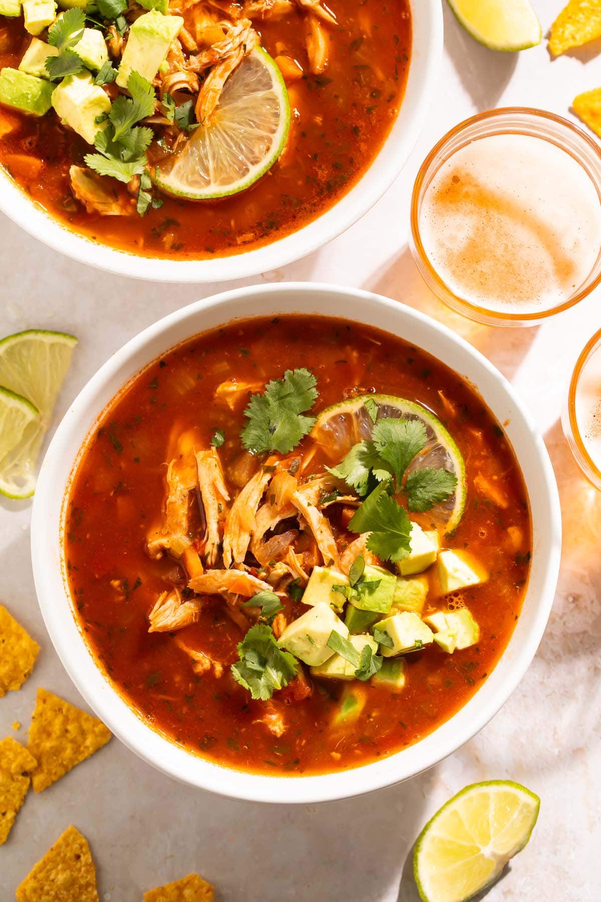 Two bowls of Mexican chicken soup on a table, surrounded by lime wedges, tortillas, and glasses of beer.