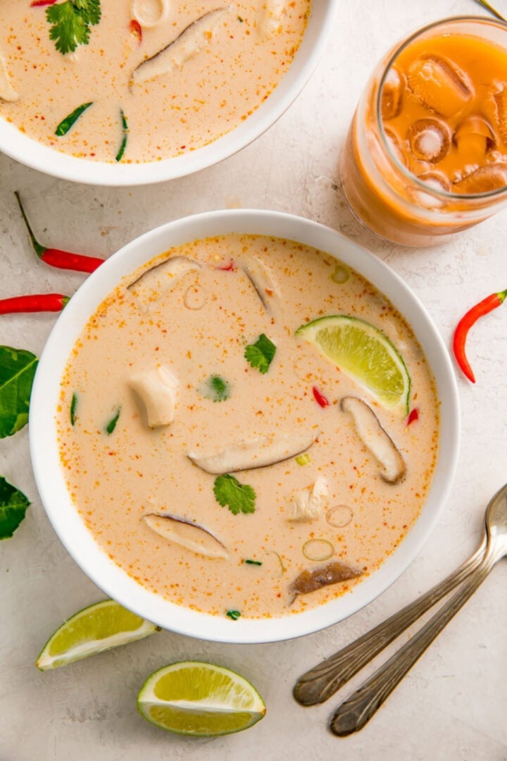 Crockpot Tom Kha Gai soup in a white bowl on a table next to lime wedges and an orange beverage in a glass.