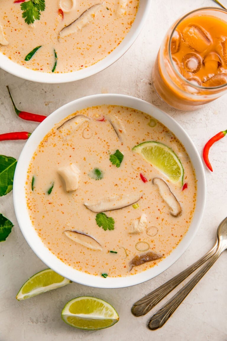 Crockpot Tom Kha Gai soup in a white bowl on a table next to lime wedges and an orange beverage in a glass.