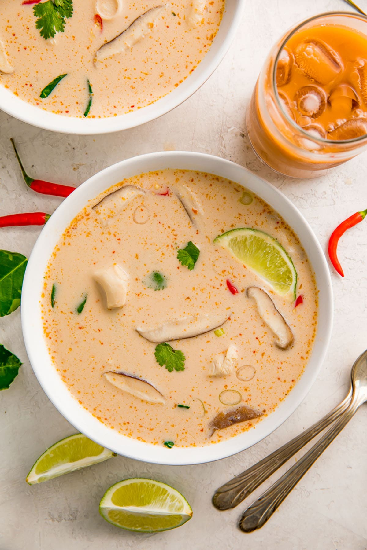 Crockpot Tom Kha Gai soup in a white bowl on a table next to lime wedges and an orange beverage in a glass.