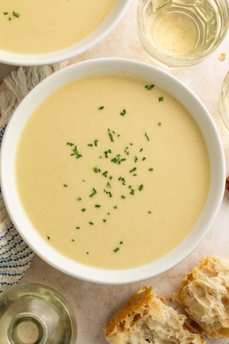 Close-up of creamy celery soup garnished with chives in a white bowl, with bread and wine nearby.