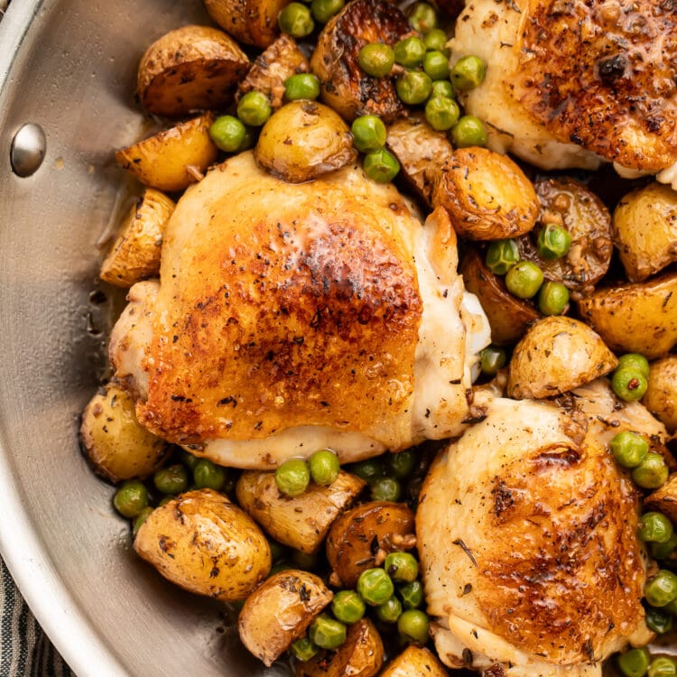 Chicken vesuvio with potatoes and peas in a large silver skillet resting on a striped kitchen towel.