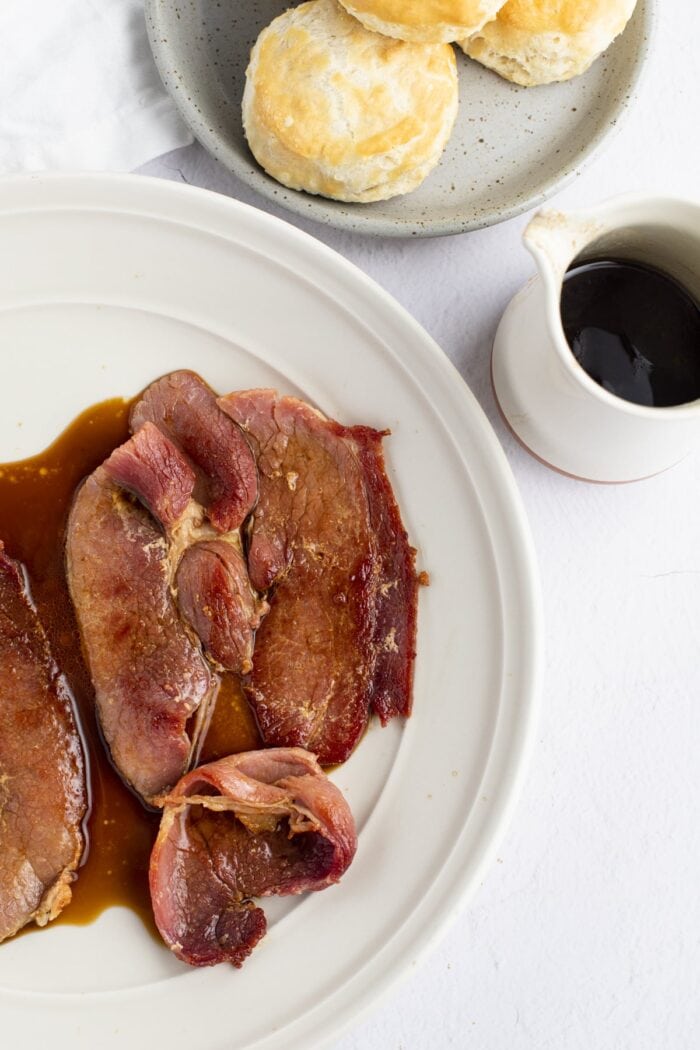 Overhead view of country ham on a white plate on the left side of the photo, with a jar of red eye gravy and a bowl of biscuits above and to the right